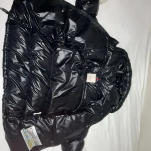Svart Moncler pufferjacka - Säljer en svart, glansig pufferjacka från Moncler med huva och dragkedja. Jackan har en klassisk Moncler-logga på bröstfickan och quiltad design. Perfekt för kalla dagar och riktigt snygg streetstil.