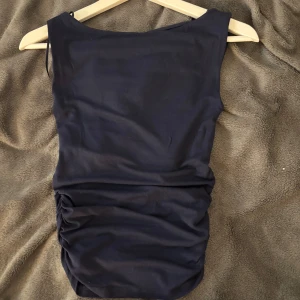 Marinblå topp - Säljer en marinblå topp från Gina Tricot i storlek XS. Toppen är ärmlös med en tight passform och har snygga rynkade detaljer på sidorna. Perfekt för dig som gillar enkel men stilren design.