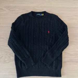 Svart kabelstickad tröja Polo Ralph Lauren - Svart kabelstickad tröja från Polo Ralph Lauren med klassisk röd logobrodyr på bröstet. Rund hals och ribbade muddar vid ärmslut och nederkant. Perfekt för dig som gillar stilren och tidlös design med en sportig touch.