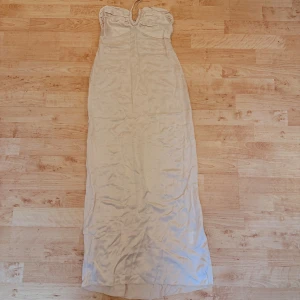 Krämfärgad långklänning från Àvora - Supersnygg långklänning från Àvora i krämfärgad ton. Klänningen har axelbandslös design med snygg rynkning upptill och knyt i nacken. Materialet känns mjukt och har ett lätt satinliknande skimmer. Perfekt för dig som vill ha en elegant och stilren look.