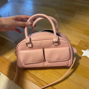 Ljusrosa handväska med dubbla fickor - Söt ljusrosa handväska i syntetmaterial med två praktiska fickor framtill och justerbar axelrem. Väskan har rundad form, dubbla handtag och dragkedja upptill. Perfekt storlek för det viktigaste och en stilren detalj till din outfit.