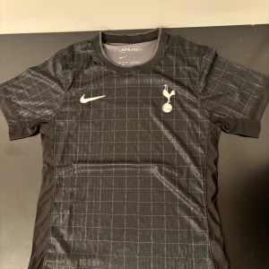 Tottenham Hotspur svart matchtröja Nike - Snygg svart Tottenham Hotspur fotbollströja från Nike med rutigt mönster och Spurs-logga på bröstet. Tröjan är kortärmad och tillverkad i 100% polyester med Dri-Fit ADV-teknologi för bästa komfort. Perfekt för dig som vill representera Spurs på träning eller match.