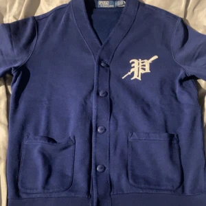 Blå kofta från Polo Ralph Lauren - Snygg marinblå kofta från Polo Ralph Lauren med klassisk knappstängning och två praktiska fickor framtill. På bröstet finns ett broderat P i vitt med en cool detalj. Mjuk bomullskvalitet och ribbade muddar ger en chill och stilren look. Nypris 3000kr, använt 1 gång, pris kan diskuteras!!
