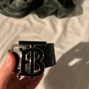 Burberry bälte med TB-spänne - Säljer ett Burberry bälte med ikoniskt TB-spänne i silverfärgad metall. Bältet har klassiskt rutigt mönster i svart och beige med inslag av brunt och grått. Tillverkat i skinn och har en stilren, modern look som passar till jeans eller chinos.