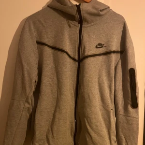 Grå Nike tech fleece  - Grå hoodiejacka från Nike med svart logga på bröstet och svarta detaljer längs dragkedjan och bröstet. Jackan har huva, lång ärm med ficka och dragkedja på ena ärmen. Perfekt för chill eller träning. Storlek XL men passar bra på L