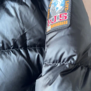 Svart dunjacka Parajumpers XS - Snygg svart dunjacka från Parajumpers, modell Sheen. Jackan har quiltad design, två snedställda dragkedjefickor framtill och klassisk PJS-patch på ärmen. Perfekt för kalla dagar och riktigt skön att bära.
