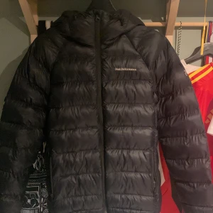 Svart dunjacka Peak Performance S - Svart dunjacka från Peak Performance i storlek S. Jackan har huva, dragkedja framtill och broderad logga på bröstet. Klassisk pufferstil med quiltade paneler och snygg, sportig look. Perfekt för kalla dagar när du vill hålla värmen och ändå se stilren ut.