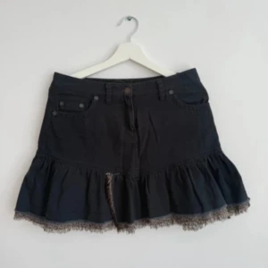 Svart volangkjol med spetskant - Supersöt svart kortkjol med volang och spetskant nertill. Kjolen har klassiska jeansdetaljer som fickor och bälteshällor, samt en liten slits framtill. Perfekt för dig som vill ha en edgy men feminin look.