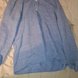 Blå linneskjorta från NN07 - Snygg blå henley-skjorta från NN07 i luftigt linne. Skjortan har lång ärm, knappar upptill och en avslappnad passform. Perfekt för dig som gillar stilren och enkel design med en fräsch vibe.