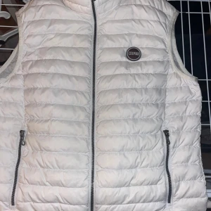 Vit dunväst från Colmar - Snygg vit dunväst från Colmar med quiltad design och dragkedja framtill. Västen har två praktiska fickor med dragkedja och en rund Colmar-logga på bröstet. Perfekt att slänga över en hoodie för en clean look. Västen sitter som storlek S.