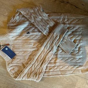 Beige kabelstickad tröja Polo Ralph Lauren - Snygg beige kabelstickad tröja från Polo Ralph Lauren med klassisk broderad logga på bröstet. Tröjan har långärmade ribbade muddar och rund hals. Perfekt för dig som gillar stilrena och tidlösa plagg med premiumkänsla.