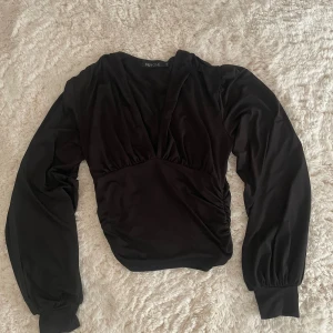Svart v-ringad blus från NLY ONE - Svart blus från NLY ONE i storlek S med djup v-ringning och långa puffärmar med breda muddar. Toppen har lätt rynkade detaljer vid midjan och axlarna för en snygg siluett. Materialet känns mjukt och har ett elegant fall.