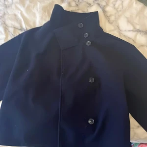 Mörkblå kappa från Gina Tricot XS - Mörkblå kappa från Gina Tricot i klassisk modell med hög krage och dubbla rader knappar framtill. Jackan har en rak passform och är perfekt för dig som gillar en tidlös look. Materialet känns mjukt och håller värmen under kyliga dagar.