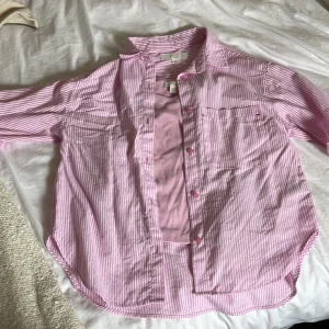 Rosa och vit randig skjorta från Zara - Snygg rosa och vit randig skjorta från Zara med klassisk krage, bröstficka och knäppning framtill. Skjortan är i bomull och har långa ärmar som kan vikas upp. Perfekt för en chill och trendig look.