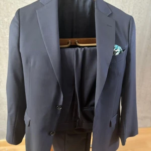 Mörkblå kostym Oscar Jacobson 48 - Snygg mörkblå kostym från Oscar Jacobson i klassisk modell. Kavajen har två knappar, spetsiga slag och en bröstficka med mönstrad näsduk. Byxorna är raka och dressade. Kostymen är tillverkad i ull och har en stilren look.