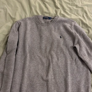 Grå stickad tröja från Polo Ralph Lauren - Snygg grå stickad tröja från Polo Ralph Lauren med klassisk rund halsringning och diskret broderad logga på bröstet. Perfekt för lager-på-lager och passar till allt. Skön och stilren med ribbade muddar vid ärmslut och hals.