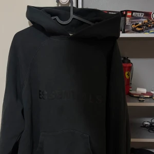 Svart Essentials Fear of God hoodie S - Svart hoodie från Essentials Fear of God i storlek S. Klassisk oversized passform med stor huva och känguruficka. Tjockt bomullsmaterial och diskret logga framtill. Perfekt för en chill och stilren streetwear-look.