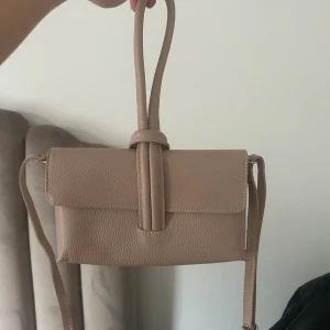 Beige/rosa handväska med knyt-detalj - Snygg beige axelväska i skinn med stilren design och unik knyt-detalj på handtaget. Väskan har en rektangulär form, lock och justerbar axelrem. Perfekt storlek för mobil, plånbok och småsaker. Enkel att matcha med olika outfits.