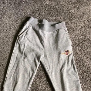 Grå mjukisbyxor från Gant - Snygga grå mjukisbyxor från Gant med broderad logga och emblem på ena benet. Byxorna har resår i midjan och sidofickor, perfekta för en avslappnad stil. Tillverkade i mjukt bomullsmaterial för extra komfort.