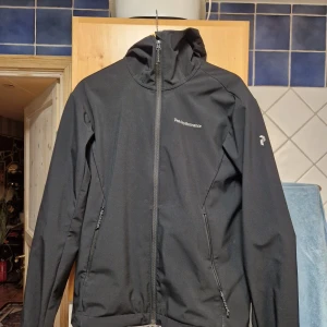 Svart vindjacka från Peak Performance - Svart vindjacka från Peak Performance med huva och hel dragkedja framtill. Jackan har två snedställda fickor med dragkedja och diskret logga på bröstet samt ärmen. Perfekt för blåsiga dagar och snygg, minimalistisk look.
