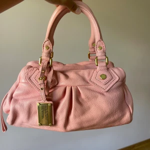 Rosa handväska från Marc by Marc Jacobs - Superfin rosa handväska i skinn från Marc by Marc Jacobs med guldfärgade metalldetaljer och dubbla handtag. Väskan har en snygg plisserad design framtill och en metallbricka med logga. Perfekt storlek för det viktigaste och ger en söt touch till din outfit.