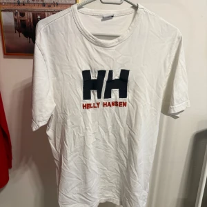 Vit Helly Hansen t-shirt med logga - Vit t-shirt från Helly Hansen med stor svart HH-logga och röd text broderad på bröstet. Klassisk rund halsringning och korta ärmar. Tillverkad i mjuk bomull för en skön känsla. Perfekt för en clean och sportig stil.