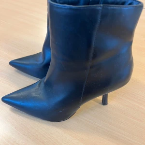 Svarta stövletter med spetsig tå - Säljer ett par svarta stövletter med spetsig tå och smal hög klack. Skorna har dragkedja på sidan för enkel på- och avtagning. Tillverkade i slätt skinnliknande material som ger en snygg och stilren look. Perfekta för dig som gillar edgy och trendiga boots.