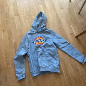 Grå Dickies hoodie med logga - Säljer en grå hoodie från Dickies med stor färgglad logga på bröstet. Tröjan har huva, magficka och långa ärmar. Perfekt för dig som gillar streetwear och vill ha en klassisk look.
