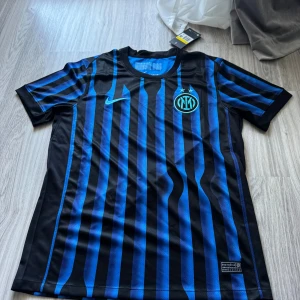Inter Milan hemmaställ 25/26 (Rabatt) - Inter Milan matchtröja från Nike i svart och blått med vertikala ränder. Klassisk rund hals, korta ärmar och klubbmärke på bröstet. Tillverkad i lätt Dri-FIT polyester som andas, perfekt för fotbollsplanen eller läktaren.