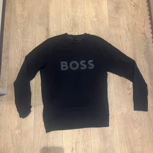 Svart sweatshirt från BOSS - Svart sweatshirt från BOSS med stor logga framtill. Tröjan har rund halsringning, långa ärmar och ribbade muddar vid ärmslut och nederkant. Tillverkad i mjuk bomull. INGA DIFFEKTER nästan som helt ny. 