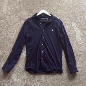 Mörkblå skjorta från Polo Ralph Lauren - Klassisk mörkblå skjorta från Polo Ralph Lauren med vit broderad logga på bröstet. Skjortan har lång ärm, knappar framtill och är tillverkad i mjuk bomull. Perfekt för en clean och stilren look. Pris kan diskuteras