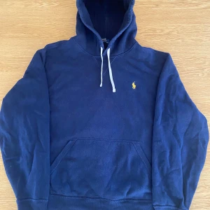 Mörkblå hoodie från Polo Ralph Lauren - Mörkblå hoodie från Polo Ralph Lauren med klassisk gul broderad logga på bröstet. Tröjan har huva med vita snören och en stor magficka framtill. Perfekt för dig som gillar stilren och sportig look.