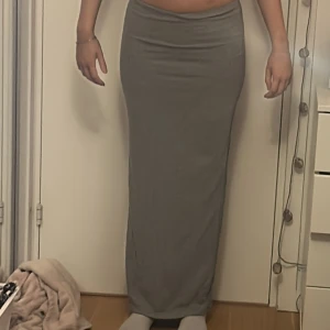 Grå långkjol i stretchigt tyg - Säljer en enkel och stilren grå långkjol med tight passform. Kjolen är rak i modellen och har en mjuk, stretchig känsla som sitter skönt på kroppen. Perfekt för dig som gillar minimalistisk stil och vill ha något bekvämt men ändå snyggt.