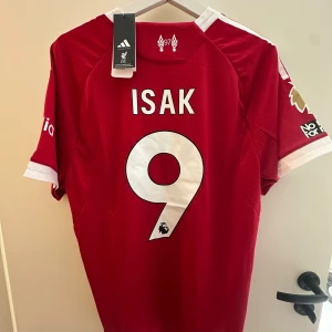 Isak Liverpool Hemmaställ 25/26 - Liverpool FCs officiella matchtröja i rött med vita detaljer, Adidas-logga och broderat klubbmärke. Nummer 9 och namnet Isak på ryggen. Tillverkad i ventilerande funktionsmaterial, kortärmad med Premier League-märke på ärmen och autentisk licensbricka.