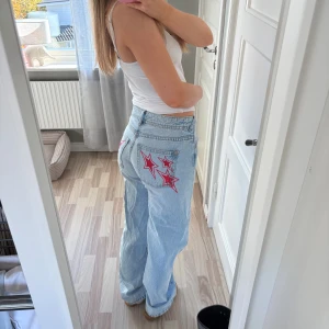Ljusblå wide jeans med stjärnor - Säljer ett par ljusblå jeans med bred passform och coola röda och rosa stjärnor broderade på bakfickorna. Jeansen har normal midja och är tillverkade i klassiskt denimtyg. Perfekta för dig som vill sticka ut lite extra.