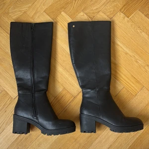 Svarta höga stövlar med klack - Snygga svarta stövlar från Din Sko skinnimitation med chunky klack och grov sula. Stövlarna har dragkedja på insidan och en stilren, minimalistisk design som passar till många outfits. Perfekta för höst och vinter när du vill ha både stil och komfort.