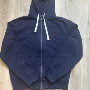 Marinblå hoodie från Polo Ralph Lauren - Marinblå hoodie från Polo Ralph Lauren med dragkedja, vit snörning i huvan och röd broderad logga på bröstet. Tröjan har långa ärmar, känguruficka och är gjord i mjuk bomull. Perfekt för en chill och stilren look. Skriv privat för mer frågor 