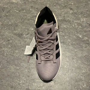 Adidas sneakers i ljuslila mesh - Snygga Adidas sneakers i grå mesh med klassiska svarta ränder på sidan. Skon har snörning och en vit och svart platt sula med coolt mönster undertill. Perfekt för dig som gillar sportig stil och vill ha något bekvämt och trendigt.