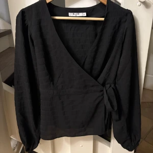 Svart omlottklänning från NA-KD - Snygg svart omlotttröja från NA-KD i storlek 34 med långa ärmar, knyt i sidan och med v-ringad hals. Tröjan är nästan aldrig använd och har inga defekter. ❣️