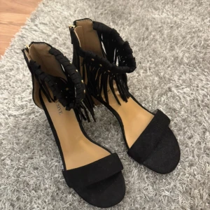 Svarta sandaler med fransar och klack - Snygga svarta sandaler från Exclusive med bred klack i trälook och öppen tå. Runt ankeln finns coola fransdetaljer och små guldfärgade nitar som ger extra edge. Materialet är mocka och sandalerna har en platt sula framtill och rund tå.