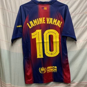 FC Barcelona Lamine Yamal tröja Nike - Säljer en officiell FC Barcelona fotbollströja från Nike med Lamine Yamal och nummer 10 på ryggen. Tröjan har klassiska blå och röda vertikala ränder, gult tryck och klubbmärke på bröstet. Materialet är lätt och andas, perfekt för match eller träning.