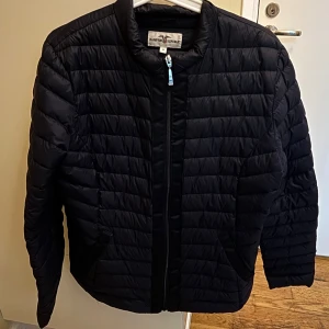 Svart pufferjacka från Hampton Republic - Svart pufferjacka från Hampton Republic med horisontella sömmar och dragkedja framtill. Jackan har en stilren, enkel design med två fickor och rundad krage. Perfekt för kyliga dagar och lätt att matcha med olika outfits.