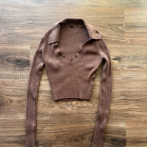  Ribbad polotop - Säljer en brun ribbad croppad polotopp med v-ringning och krage. Toppen har långa ärmar och är figurnära i modellen, perfekt för en trendig och stilren look.