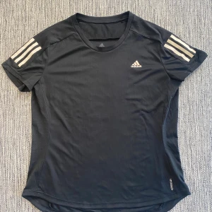 Svart Adidas t-shirt med ränder - Svart t-shirt från Adidas med klassiska vita ränder på axlarna och logga på bröstet. Gjord i lätt och ventilerande polyester, perfekt för träning. Modellen är kortärmad och har en sportig passform. AEROREADY-material som håller dig torr under aktivitet. Mått Över bröstet: ca 54 cm Längd från axeln och ner: ca 66 cm  Kommer från ett djur och rökfritt hem.