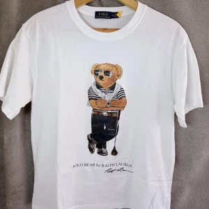Vit Polo Bear t-shirt från Ralph Lauren - Vit t-shirt från Polo Ralph Lauren med ikoniskt Polo Bear-tryck framtill. Klassisk rund hals och korta ärmar. Tillverkad i mjuk bomull för en skön känsla. Perfekt för dig som gillar stilrena plagg med en twist.