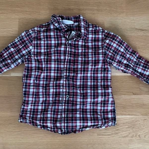 Rutig skjorta i rött och svart, stl 92 - Klassisk rutig skjorta i rött, svart och vitt med långa ärmar och knappar framtill. Skjortan har en mjuk känsla och är tillverkad i bomull, perfekt för en cool och avslappnad look.