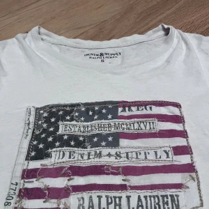 Ralph Lauren T-Shirt - En schysst Ralph Lauren T-shirt med ett skrovligt tyg tryck. Står S, men passar som en Medium. Helt okej skick, varsitt litet ihopsydd hål under armhålorna som inte syns. Nypris 1199, men jag säljer den lite billigare pga defekterna.