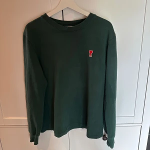 Grön långärmad tröja från AMI Paris - Mörkgrön långärmad tröja från AMI med klassisk passform och rund halsringning. På bröstet sitter ett litet rött hjärta med AMI-loggan. Tröjan är i mjuk bomull och passar perfekt för en chill och stilren look.