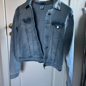 Mjuk och stretchig jeansjacka från H&M - Snygg ljusblå jeansjacka från H&M med klassisk krage, bröstfickor med knappar och silverfärgade detaljer. Jackan är tillverkad i jättestretchig denim med bomull, polyester och elastan. Snyggt tillskott till garderoben 