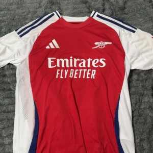 Arsenal Saka 7 matchtröja Adidas - Riktigt snygg Arsenal fotbollströja från Adidas med Saka 7 på ryggen. Tröjan är röd framtill med vita ärmar och blå detaljer, samt klubbens logga och Emirates Fly Better-tryck på bröstet. Tillverkad i lätt och ventilerande material, perfekt för match eller träning.